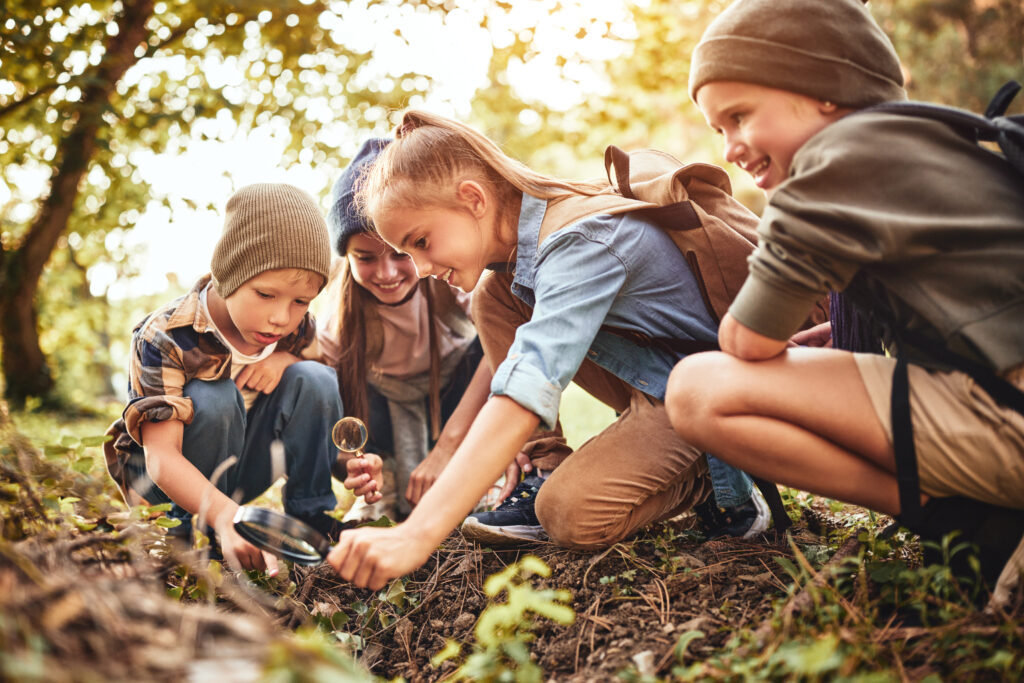 Eine Gruppe von vier Kindern erkundet neugierig den Waldboden. Sie benutzen Lupen, um kleine Pflanzen und Insekten zu untersuchen. Die Kinder tragen Rucksäcke und sind von Bäumen umgeben, was eine abenteuerliche und entdeckungsfreudige Atmosphäre vermittelt.